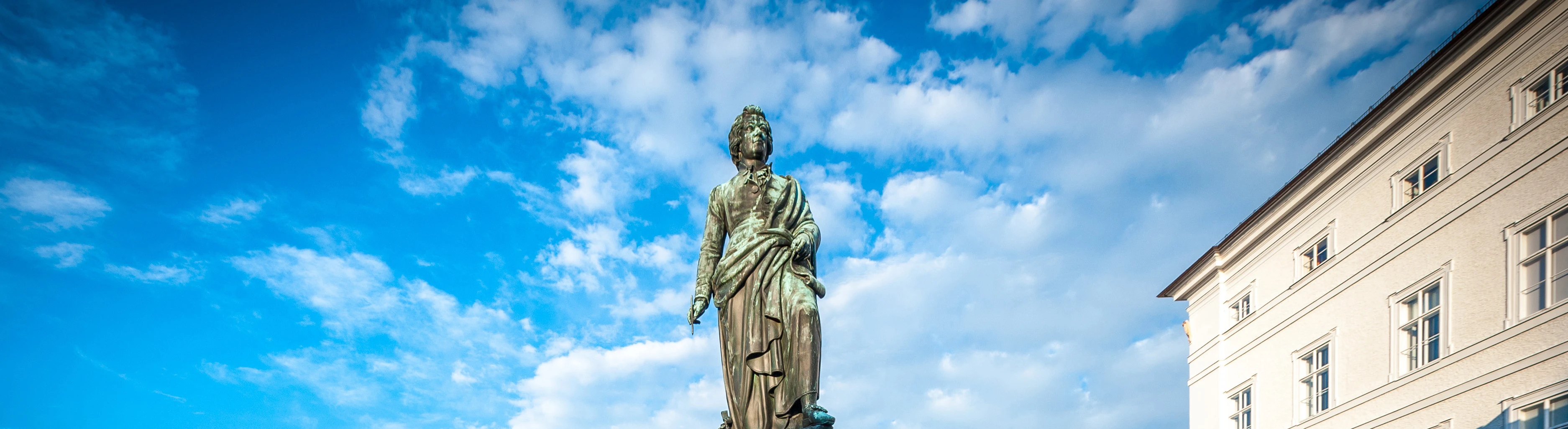The image shows the Mozart monument in Salzburg, Austria, set against a vivid blue sky with scattered clouds.