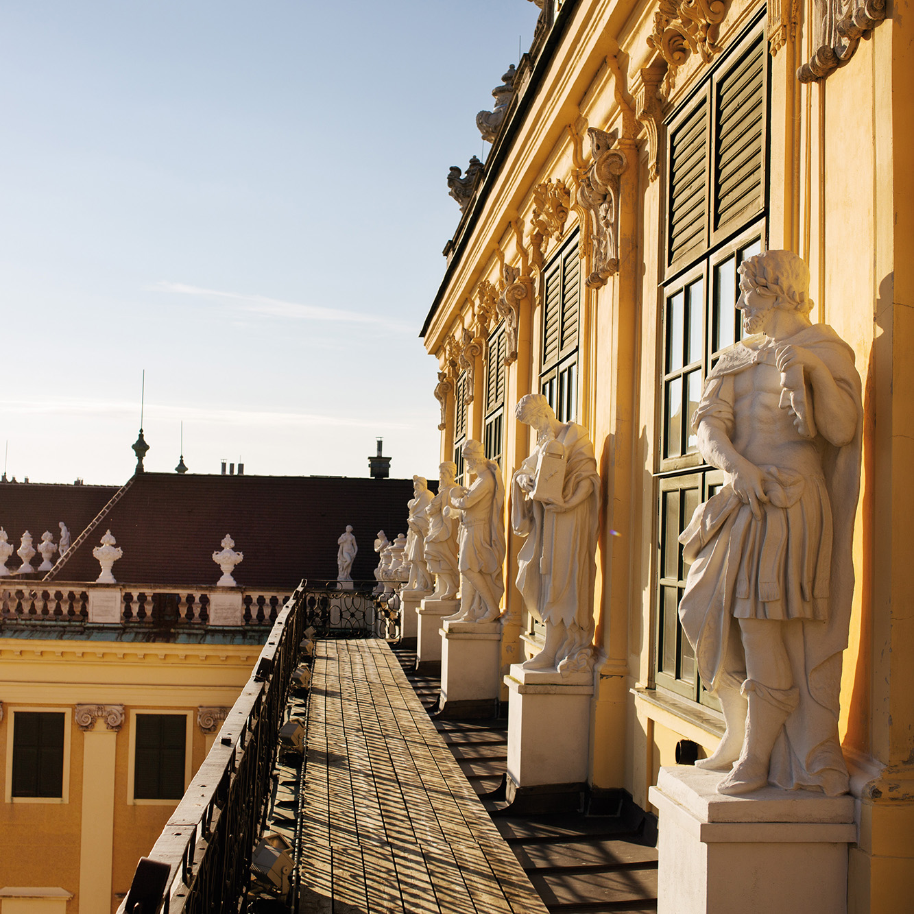 Marble statues line the sunlit balcony of Schönbrunn Palace, casting long shadows on the golden façade.