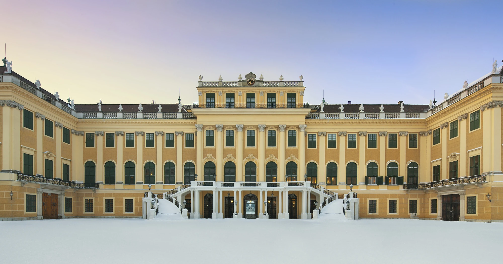 Schönbrunn Palace stands serene under a soft winter twilight, its yellow façade contrasting with the blanket of snow.