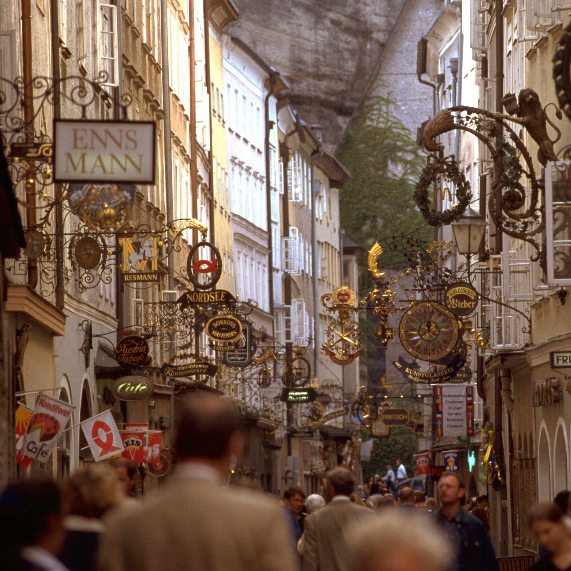 Salzburg’s Getreidegasse bursts with ornate shop signs, narrow buildings, and a view of church spires and mountains beyond.