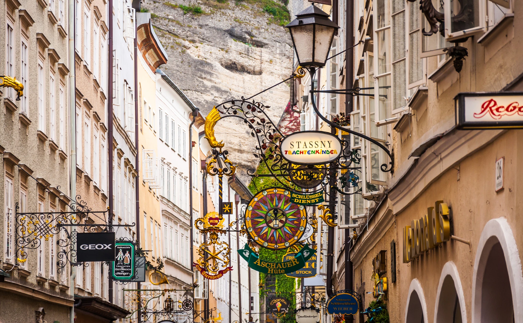 This image captures Salzburg's famous Getreidegasse, a charming historic street lined with ornate wrought-iron shop signs and flanked by old buildings set against a mountainous backdrop.