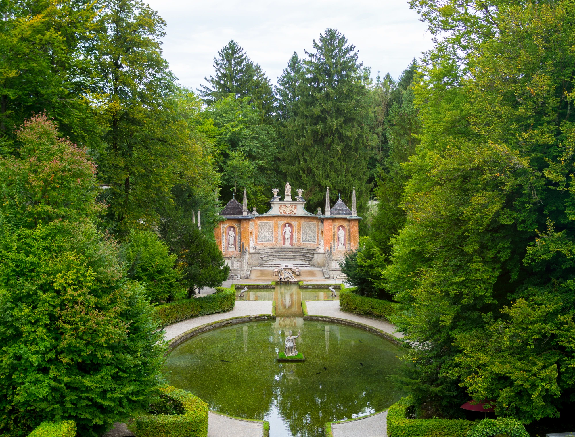 This image depicts the ornate and theatrical Roman Theatre grotto at Hellbrunn Palace, surrounded by lush forest and reflected in a circular pool with classical statues.
