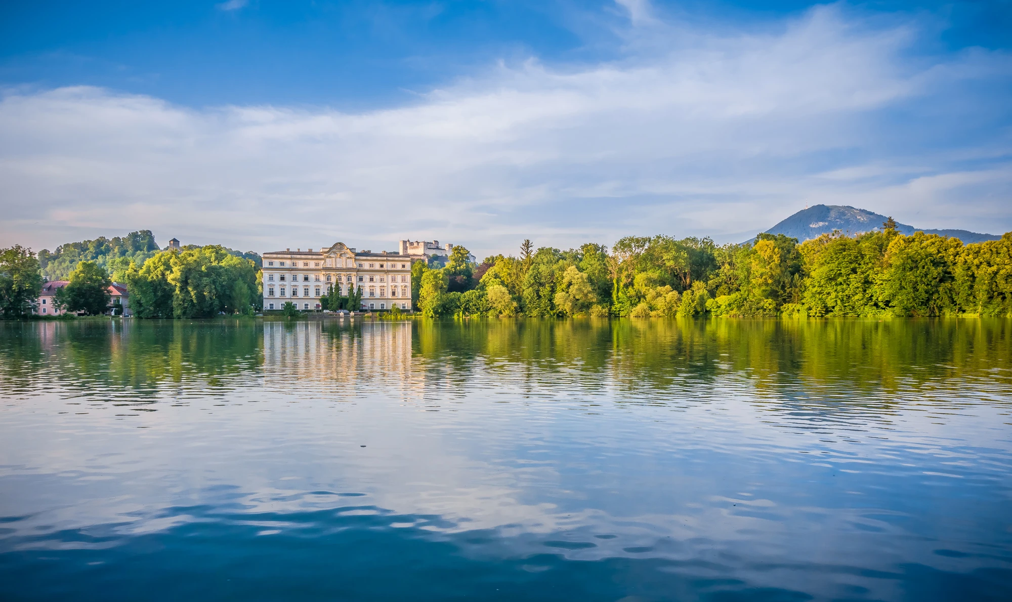 This image showcases the serene Leopoldskron Palace reflected in a calm lake, surrounded by lush greenery and backed by mountains under a vibrant blue sky.