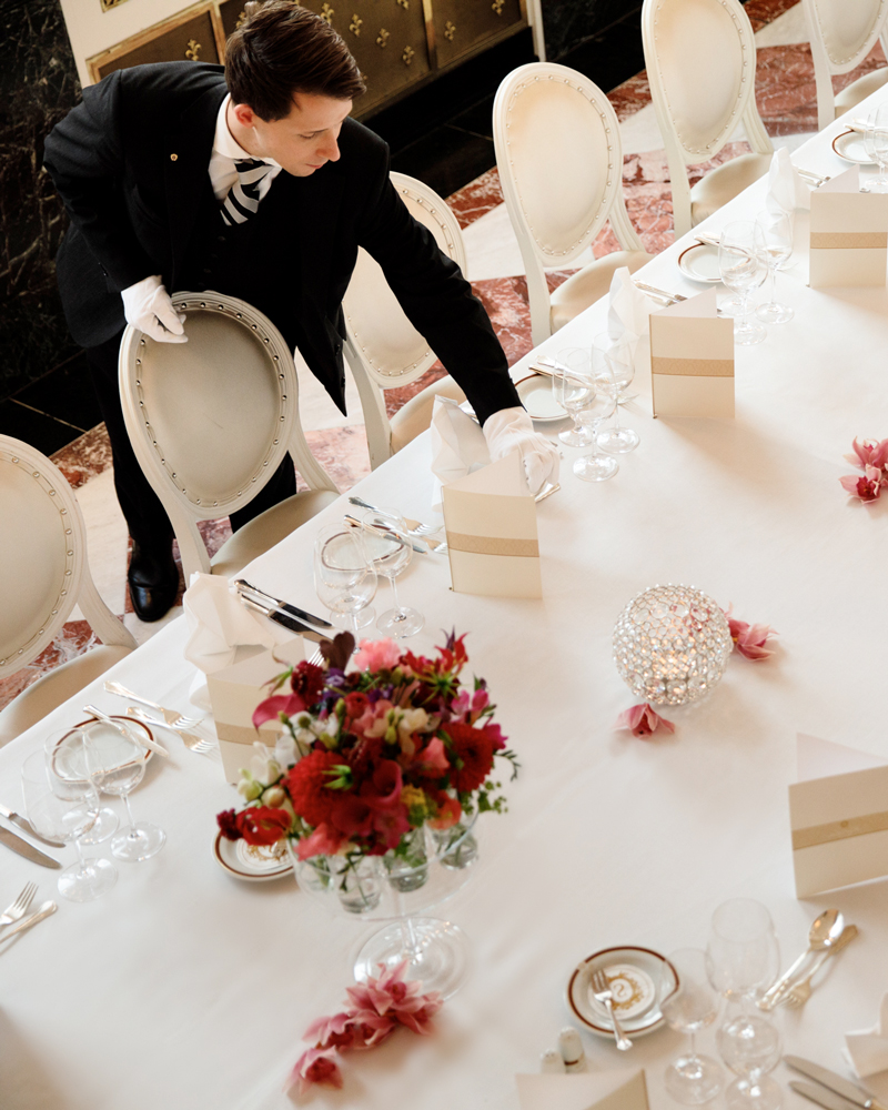 A formally dressed staff member meticulously sets an elegant table adorned with floral arrangements and fine tableware, preparing for a sophisticated dining event.