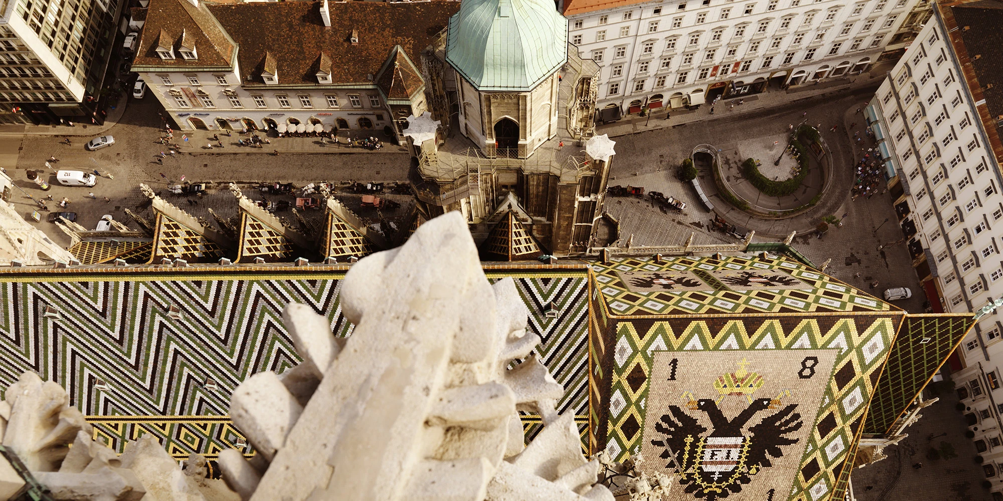The image captures a dramatic top-down view from St. Stephen’s Cathedral in Vienna, showcasing its vivid patterned roof and the lively square below.