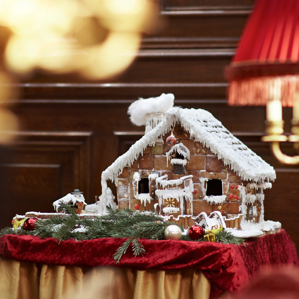 A festive gingerbread house decorated with icing and ornaments sits on a red velvet-covered table.