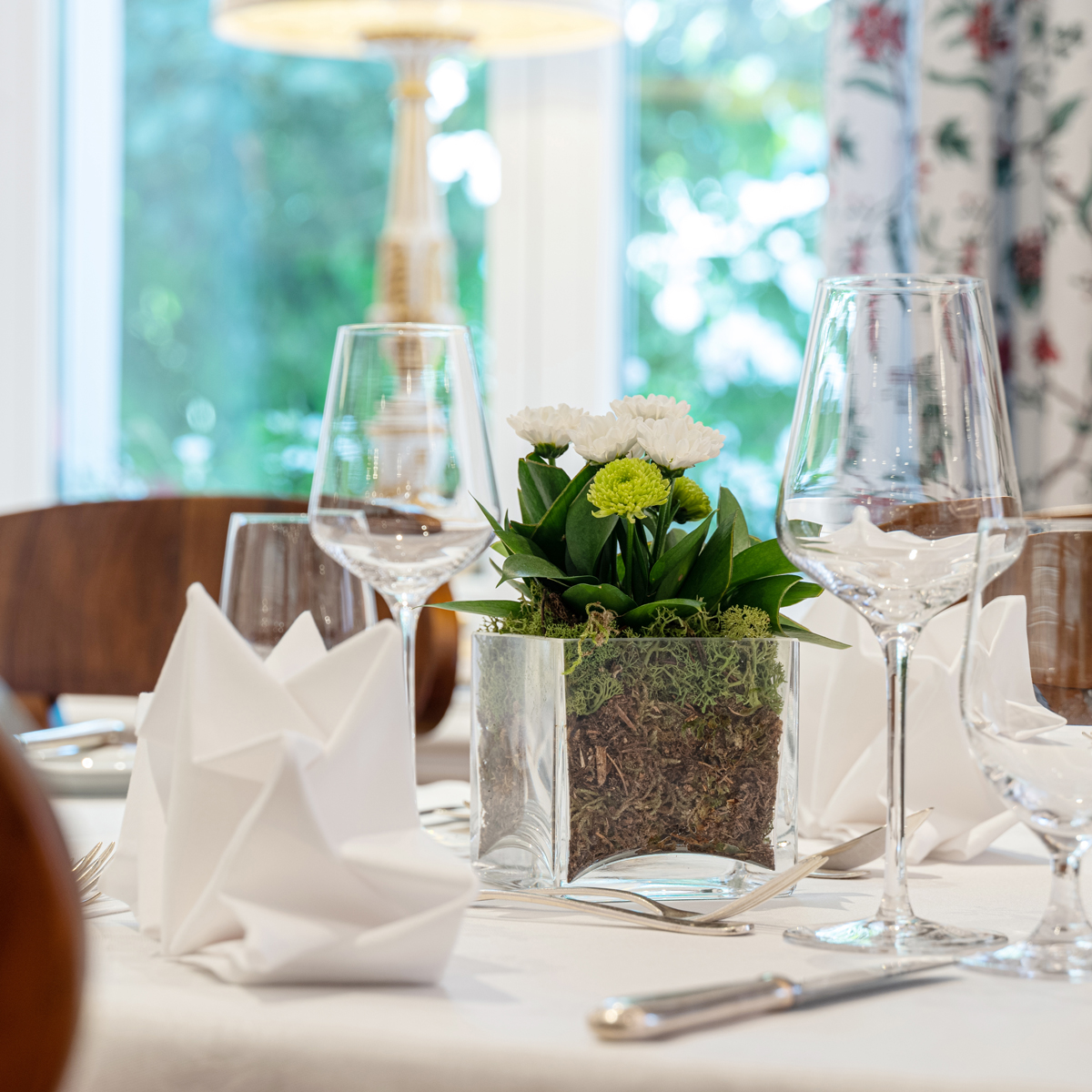 This image presents a beautifully set dining table in an elegant, light-filled room, featuring crisp white napkins, sparkling glassware, and a floral centerpiece in a transparent planter filled with moss and soil.