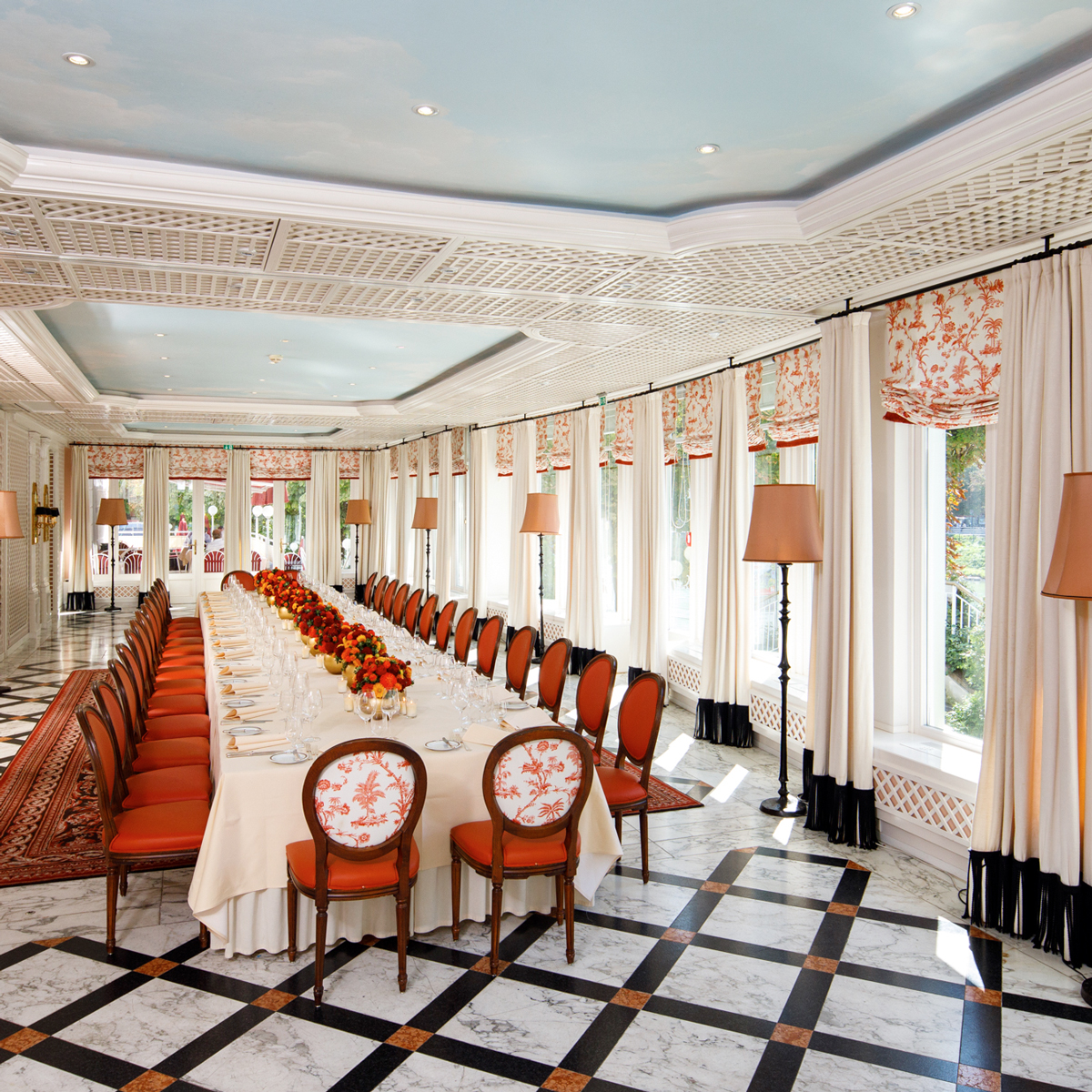 Sacher_Salzburg_Events_Meetings_Intro This image shows a long, elegant banquet table set for a formal meal, with orange chairs, floral arrangements, and large windows lining a bright, stylish dining room.