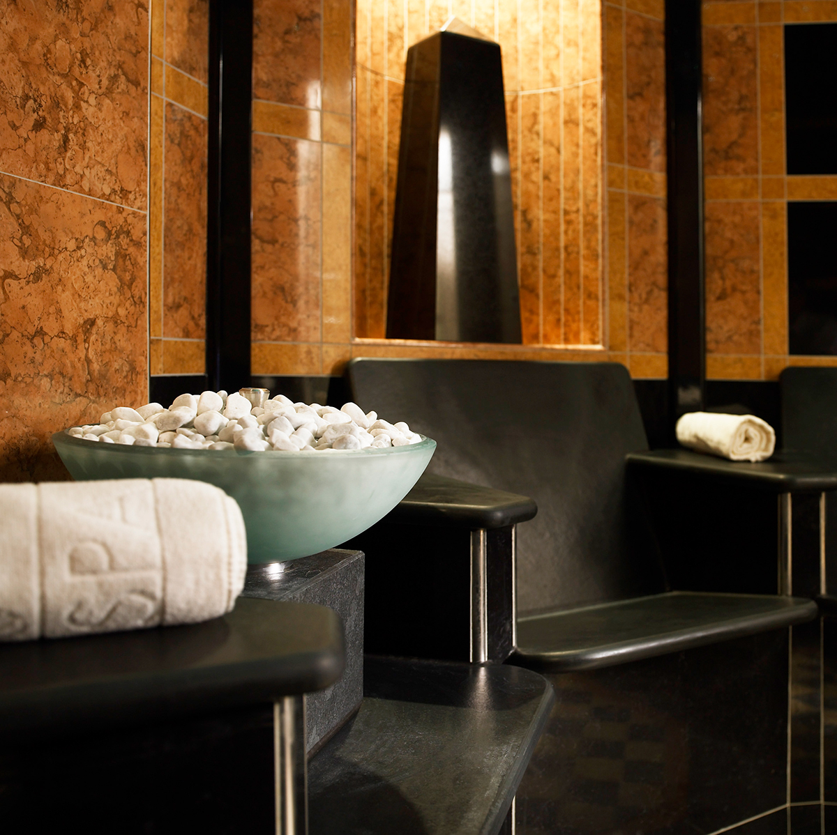 Sacher_Wien_Teaser-DaySpa-2-2 A serene spa steam room with black stone seating, a frosted bowl of white stones, and neatly rolled towels.