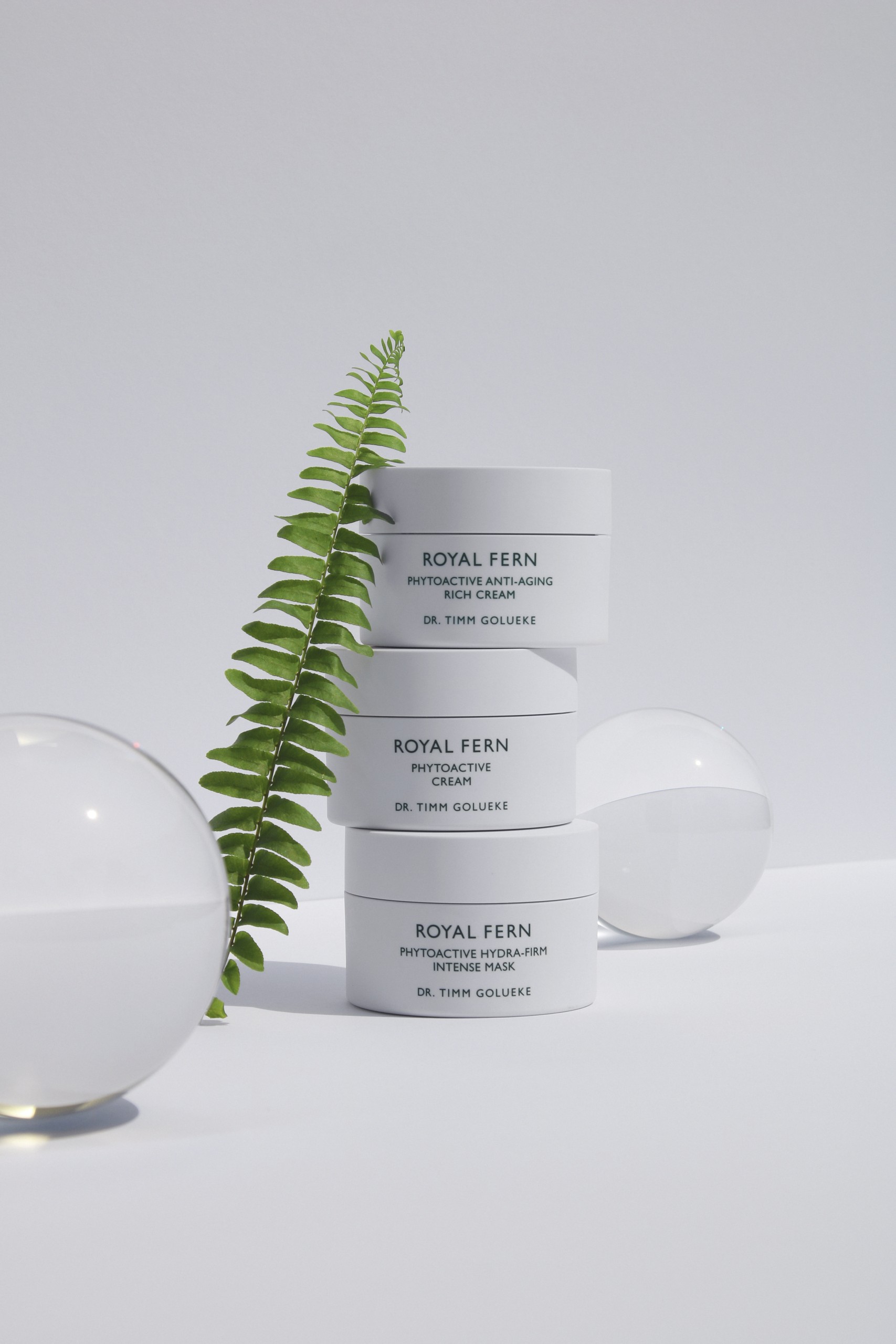 2021_06_marvinungerstyling_3-1-scaled This image features an elegant, minimalist arrangement of three skincare products from **Royal Fern by Dr. Timm Golueke**, including: - **Phytoactive Anti-Aging Rich Cream** - **Phytoactive Cream** - **Phytoactive Hydra-Firm Intense Mask** The products are stacked vertically, framed by a delicate green fern and clear spherical ornaments—an aesthetic nod to nature and purity that aligns with the brand's botanical, science-driven ethos.