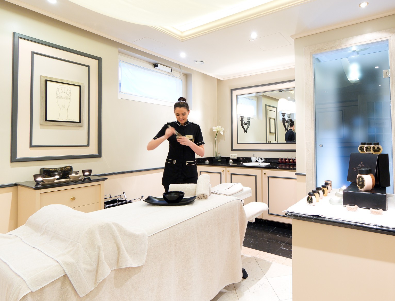 The image shows a serene treatment room at the Sacher Spa, where a spa therapist is preparing a treatment with signature products, surrounded by soft lighting, elegant décor in cream and black tones, and a neatly arranged massage bed—creating a luxurious and calming atmosphere.