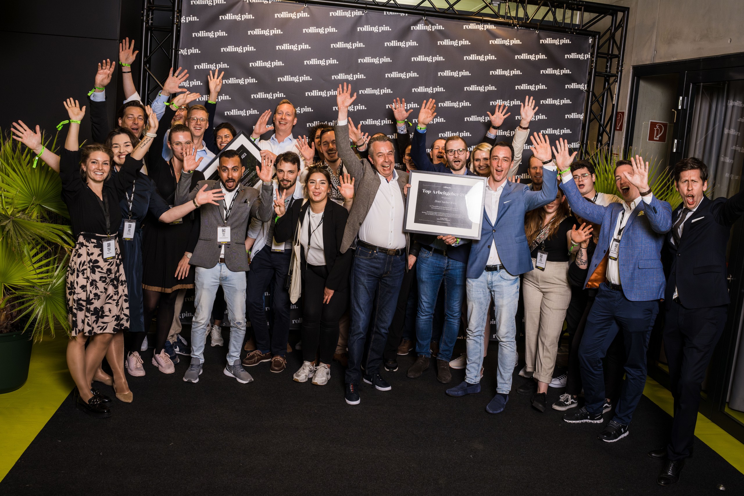 The image shows a large, joyful group of people posing together in front of a backdrop that says “rolling pin,” with many raising their hands in celebration while one man in front proudly holds a framed award labeled “Top Arbeitgeber 2024” (Top Employer 2024).