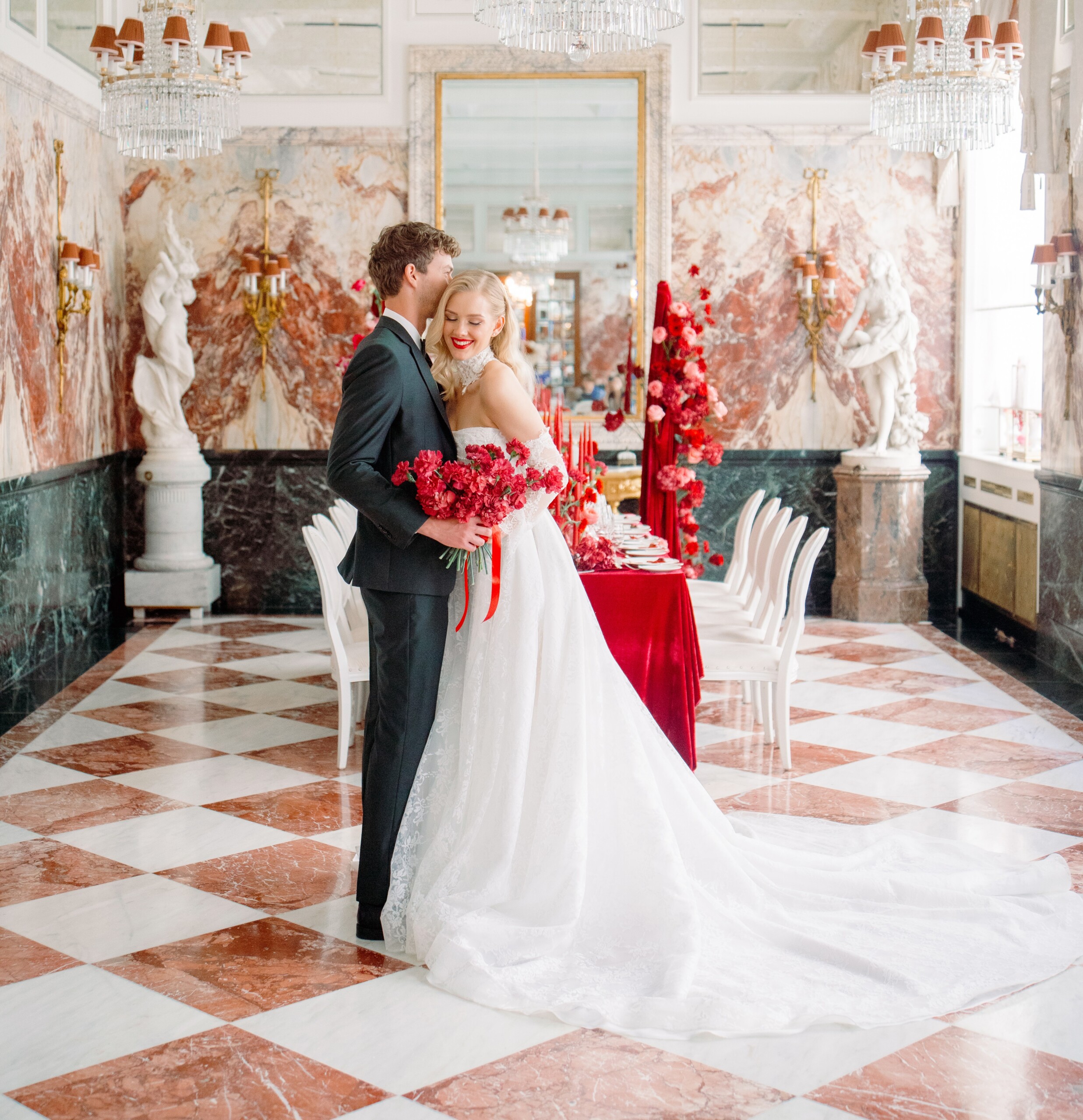 A bride and groom holding a red bouquet in front of a festively set wedding table in the Marble Hall of the Hotel Sacher Vienna.