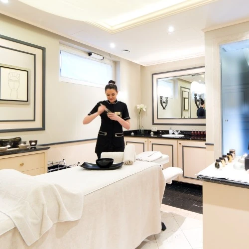 The image shows a serene treatment room at the Sacher Spa, where a spa therapist is preparing a treatment with signature products, surrounded by soft lighting, elegant décor in cream and black tones, and a neatly arranged massage bed—creating a luxurious and calming atmosphere.