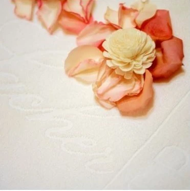 The image shows delicate rose petals and a cream-colored preserved flower arranged on a luxurious white towel embossed with the word 