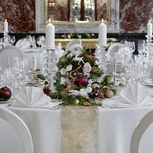 The image shows a lavish holiday setup for Christmas and New Year’s Eve in the Marmorsaal at Hotel Sacher Wien, featuring elegantly set round tables with white linens, crystal candleholders, and festive centerpieces adorned with pinecones, ornaments, dried fruits, and delicate florals, all in a grand marble-lined setting.