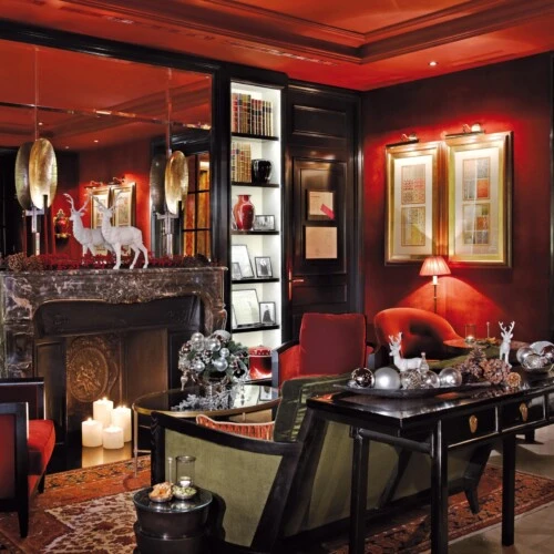 luxurious festively decorated lounge at Hotel Sacher, featuring deep red walls, elegant dark wood furniture, a grand marble fireplace adorned with white deer figurines and holiday ornaments, and cozy seating illuminated by warm, ambient lighting.