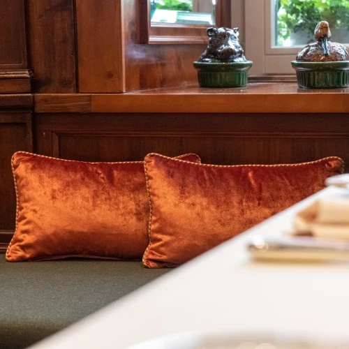 The image shows a cozy corner of the Zirbelzimmer, featuring warm wooden paneling, plush orange velvet cushions, checkered pillows, and charming ceramic animal decor on the windowsill—creating a rustic yet refined alpine atmosphere.