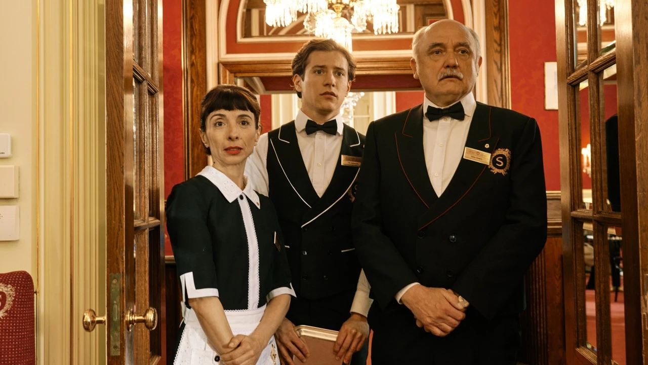 The image is a scene from the Amazon Prime series Sachertorte, showing three Hotel Sacher staff members—two men and one woman—in formal uniforms standing in a richly decorated red and gold interior, looking on with serious expressions, highlighting the drama and elegance of the hotel's atmosphere.