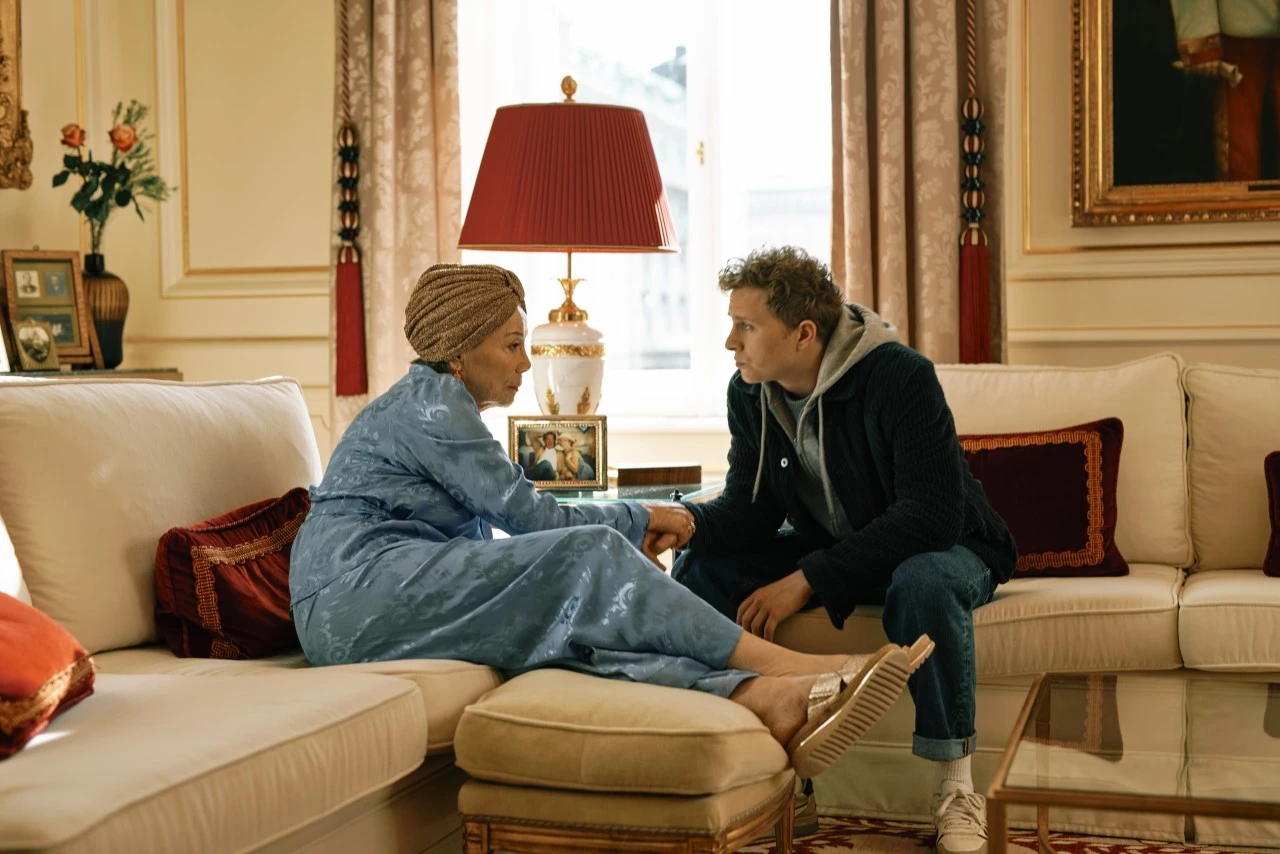 The image is another scene from the Amazon Prime series Sachertorte, showing an emotionally charged conversation between two characters in an opulently furnished hotel suite, with golden accents, rich red details, and classic décor that evokes the elegance of Hotel Sacher.