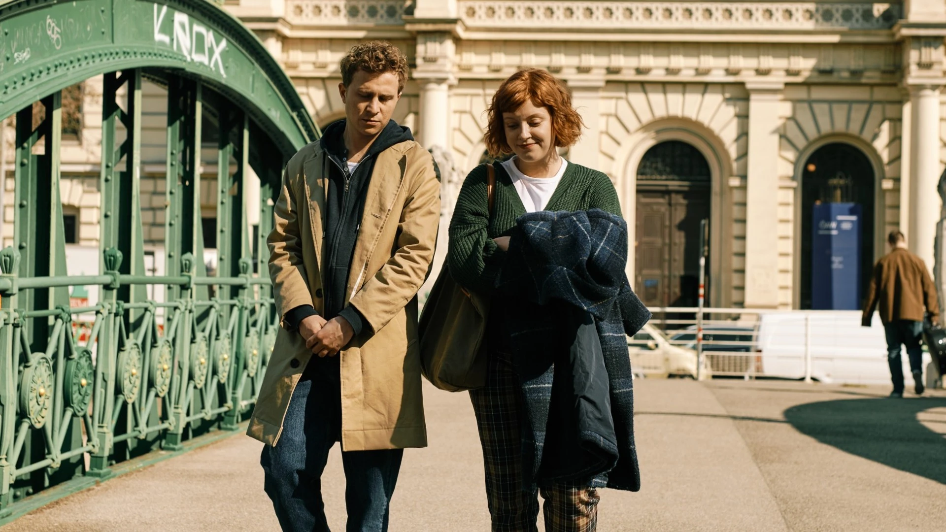 The image is a scene from the Amazon Prime series Sachertorte, showing two characters walking pensively across a historic green iron bridge in Vienna, with classical architecture in the background, capturing a quiet, emotional moment.