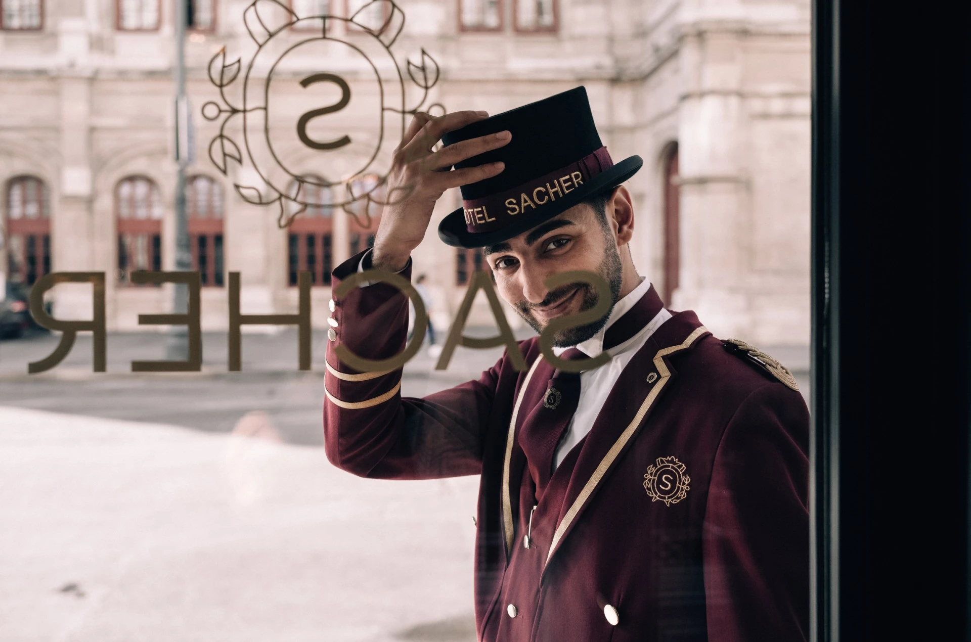 Hotel Sacher doorman tipping his hat with a warm smile, standing in front of the iconic Vienna hotel entrance.