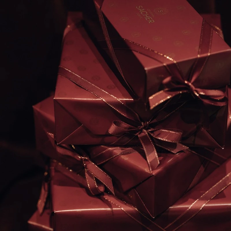 A luxurious stack of burgundy-wrapped Original Sacher-Torte boxes rests elegantly on a velvet-covered chair, ready to be gifted from Hotel Sacher Wien.