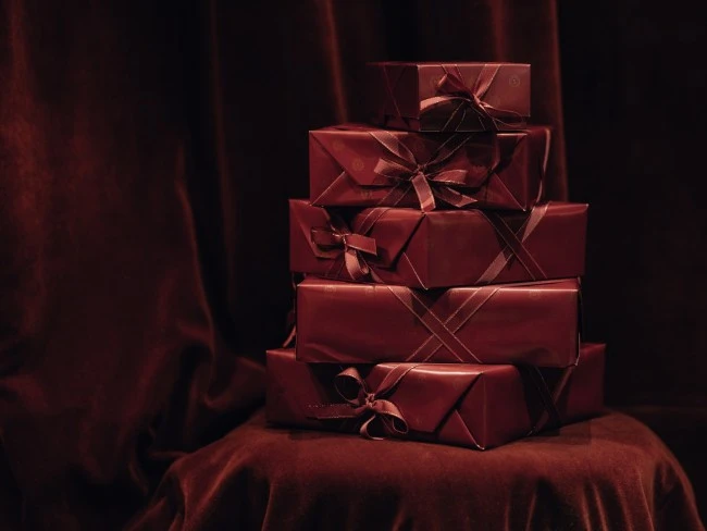 A luxurious stack of burgundy-wrapped Original Sacher-Torte boxes rests elegantly on a velvet-covered chair, ready to be gifted from Hotel Sacher Wien.