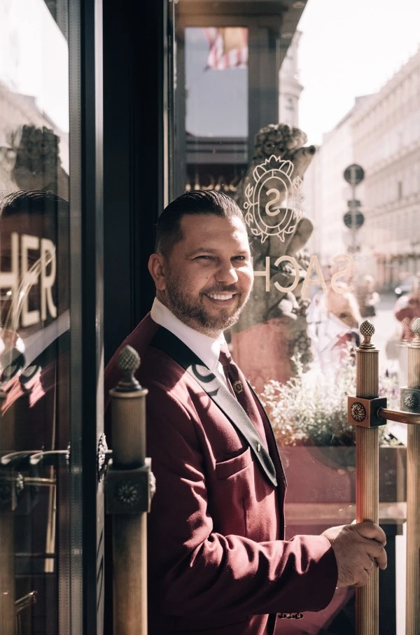 A smiling doorman in a burgundy uniform warmly greets guests at the elegant entrance of Hotel Sacher Wien.