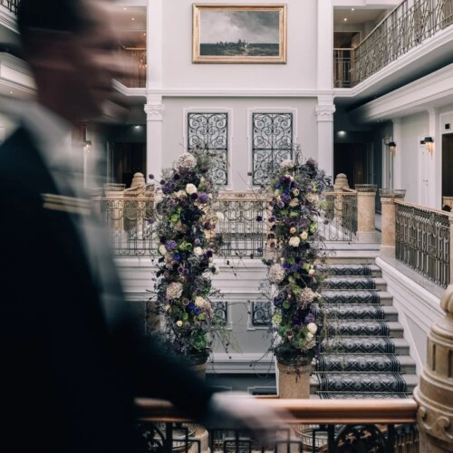 A blurred concierge walks past the grand staircase of Hotel Sacher Salzburg, which is beautifully decorated with towering floral arrangements and surrounded by elegant iron railings and classical architecture.