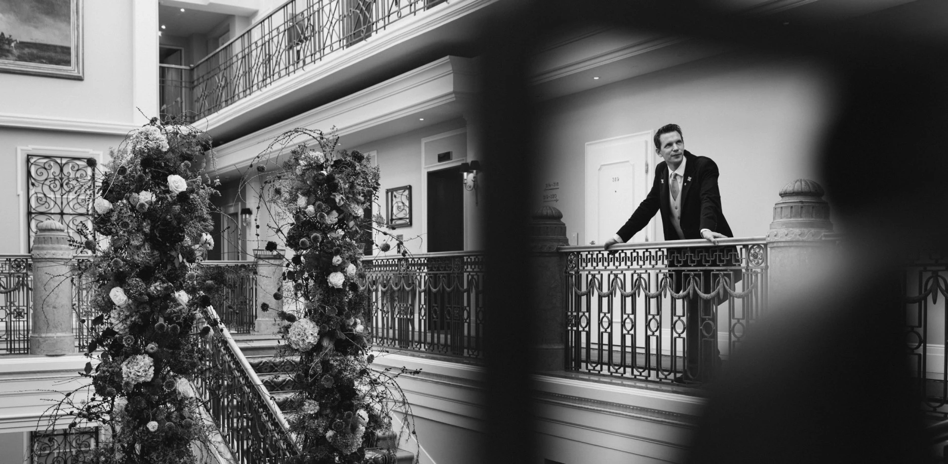 A black-and-white photo of a Hotel Sacher Salzburg concierge standing on an upper floor balcony, overlooking a staircase adorned with lush floral arrangements.