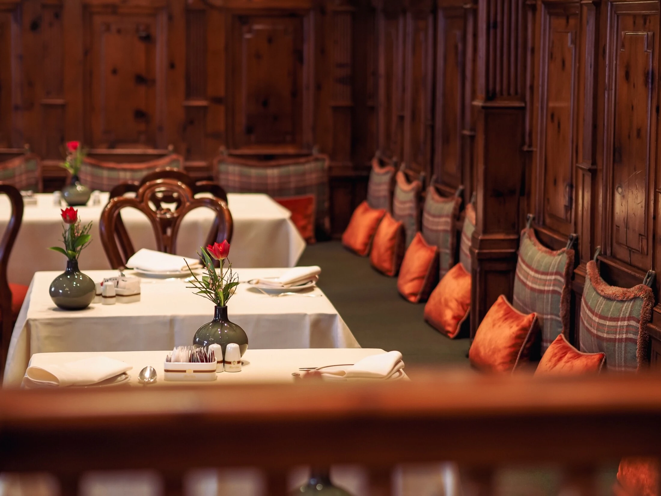 A cozy and elegant setting in the Zirbelzimmer at Hotel Sacher Salzburg, featuring wooden paneled walls, white tablecloth-covered tables, floral centerpieces, and cushioned bench seating with plaid and orange pillows.