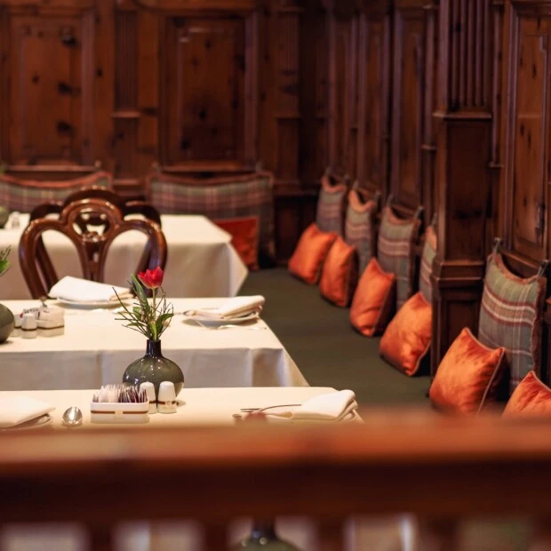 A cozy and elegant setting in the Zirbelzimmer at Hotel Sacher Salzburg, featuring wooden paneled walls, white tablecloth-covered tables, floral centerpieces, and cushioned bench seating with plaid and orange pillows.