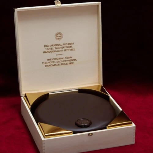 The Original Sacher-Torte is elegantly presented in a wooden box with gold corners, resting on a dark marble table in front of a richly patterned red sofa with decorative pillows.