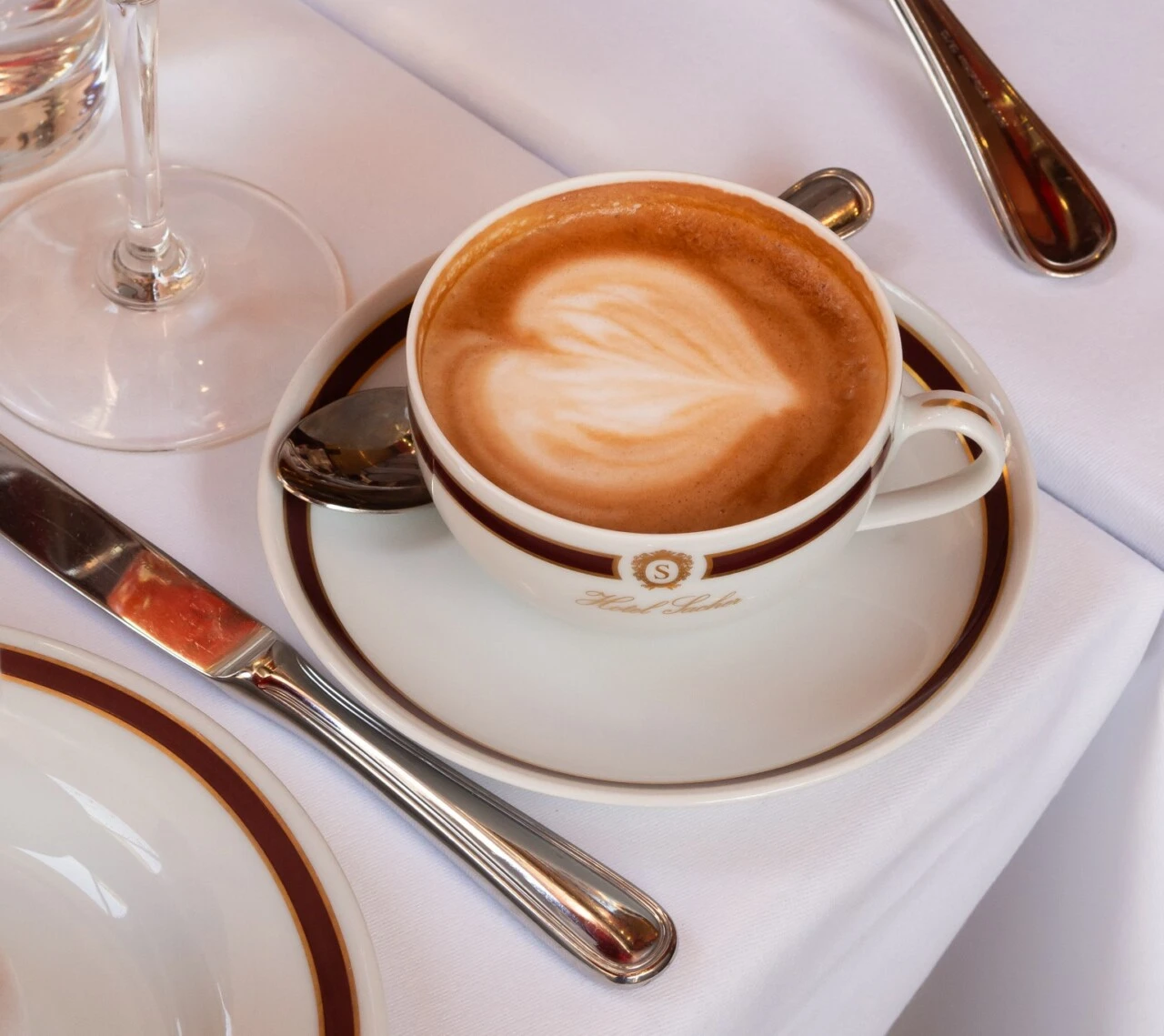 A cappuccino with heart-shaped latte art served in a Hotel Sacher cup on a white tablecloth with elegant silverware.