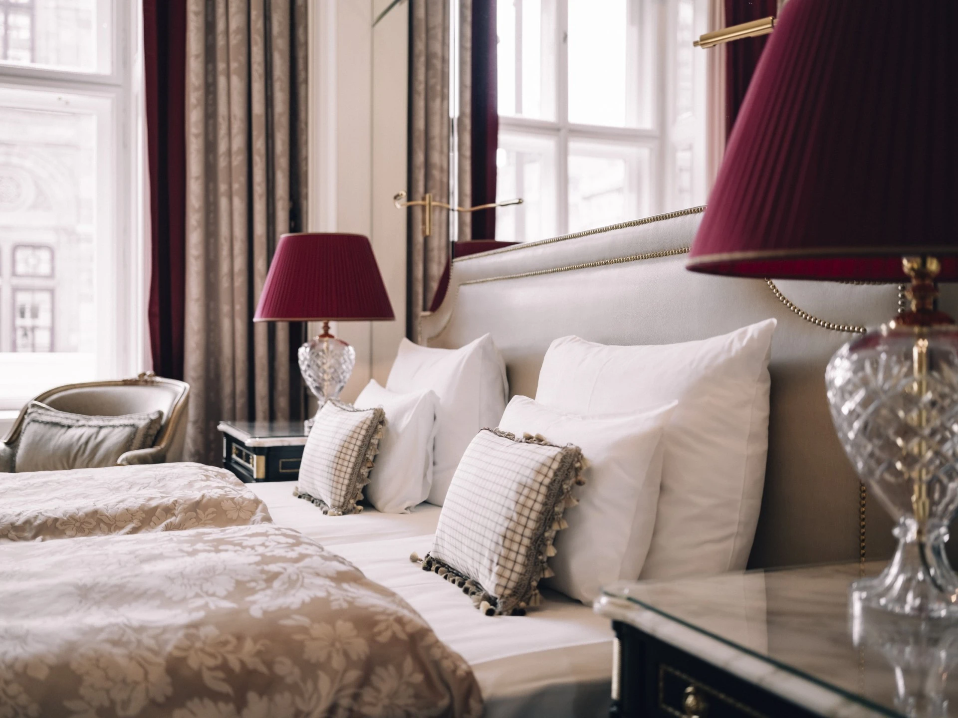 A luxurious hotel room featuring twin beds with elegant bedding, plush cushions, crystal lamps with red shades, and tall windows framed by patterned drapes.