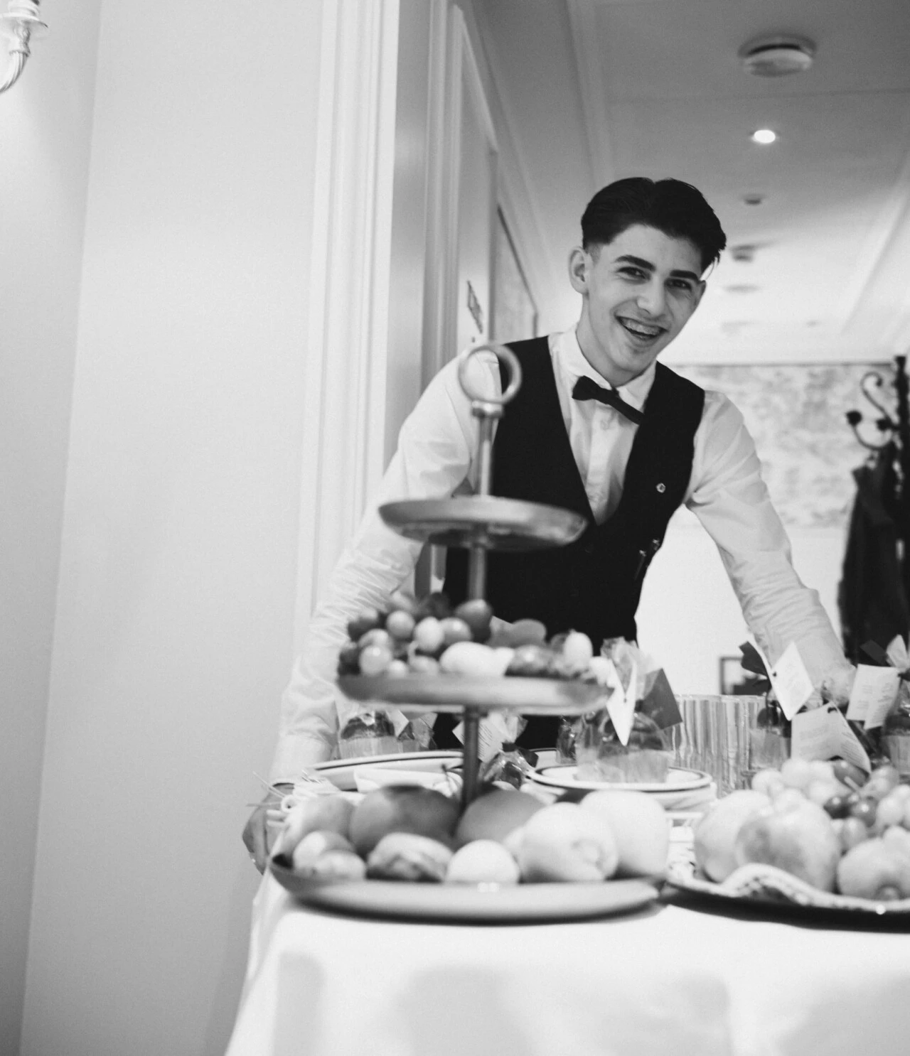 A smiling waiter in a bow tie and vest stands behind a tiered tray of fruit and pastries in a bright, elegant dining area.