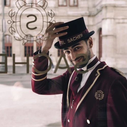 Hotel Sacher doorman tipping his hat with a warm smile, standing in front of the iconic Vienna hotel entrance.