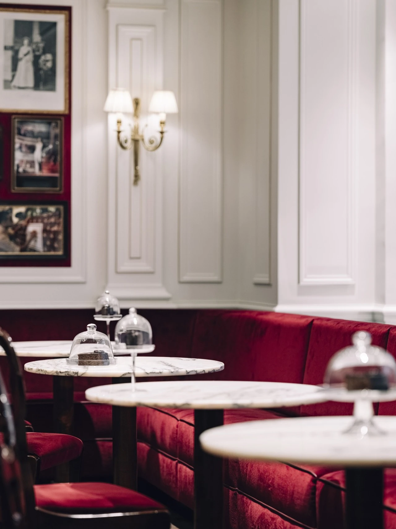 A cozy corner of Café Sacher in Salzburg featuring red velvet seating, white paneled walls with framed photos, and marble tables displaying slices of Original Sacher-Torte under glass domes.