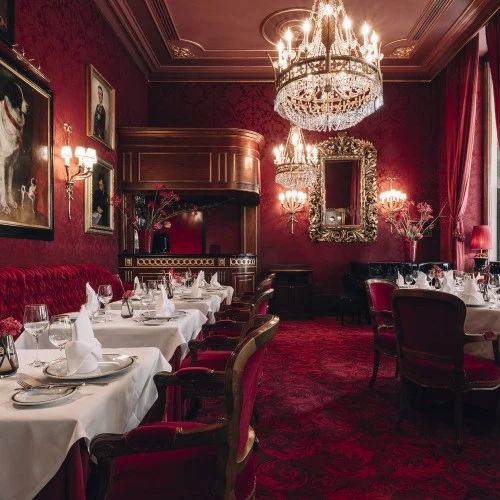 The opulent dining room of Restaurant Rote Bar features crystal chandeliers, red velvet furnishings, white tablecloths, and classical paintings on richly patterned red walls.