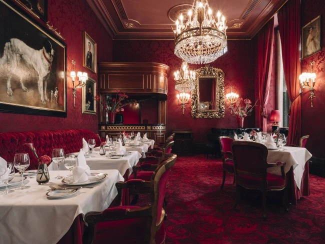 The opulent dining room of Restaurant Rote Bar features crystal chandeliers, red velvet furnishings, white tablecloths, and classical paintings on richly patterned red walls.