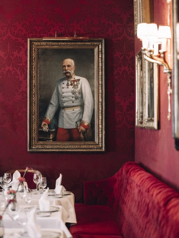 A richly decorated dining area in Restaurant Rote Bar featuring a formal table setting, plush red seating, and a large portrait of an Austrian officer in a white uniform with medals on a patterned red wall.