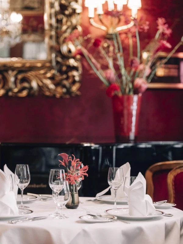 A formally set dining table with white linens and glassware in Restaurant Rote Bar, surrounded by deep red walls, ornate gold-framed mirrors, and floral arrangements.