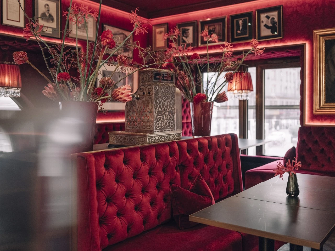 A richly decorated seating area in Restaurant Bel Etage with plush red velvet booths, floral arrangements, framed portraits, and a vintage cash register as a centerpiece.