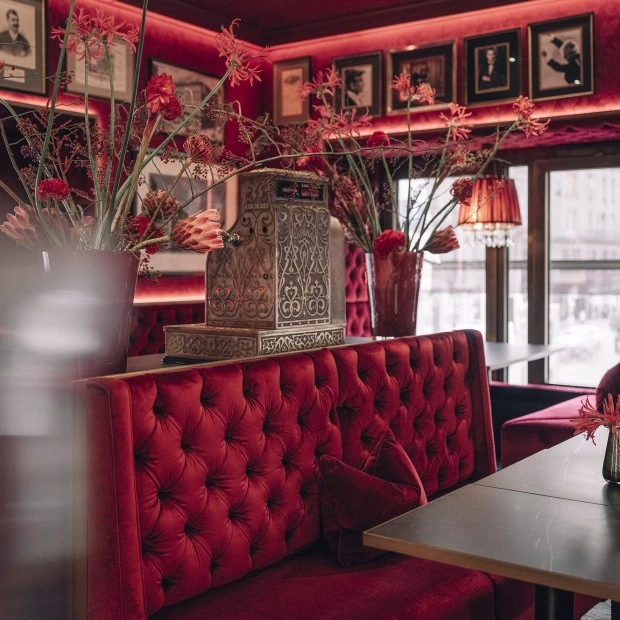 A richly decorated seating area in Restaurant Bel Etage with plush red velvet booths, floral arrangements, framed portraits, and a vintage cash register as a centerpiece.