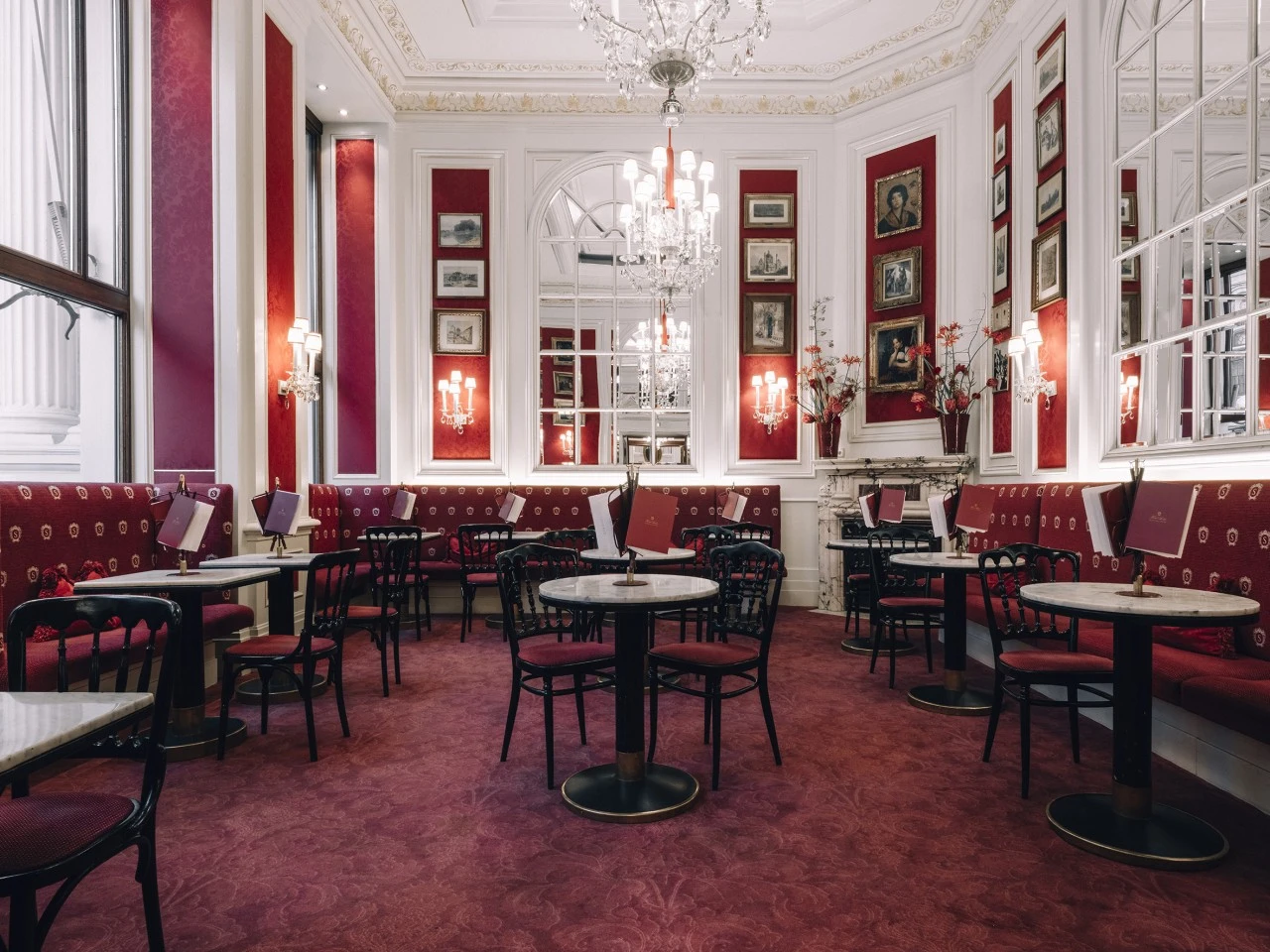 The image shows a lavish, old-world café interior with red velvet seating, marble tables, crystal chandeliers, and classic artwork, capturing the timeless elegance of Café Sacher Wien.