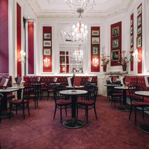 The image shows a lavish, old-world café interior with red velvet seating, marble tables, crystal chandeliers, and classic artwork, capturing the timeless elegance of Café Sacher Wien.