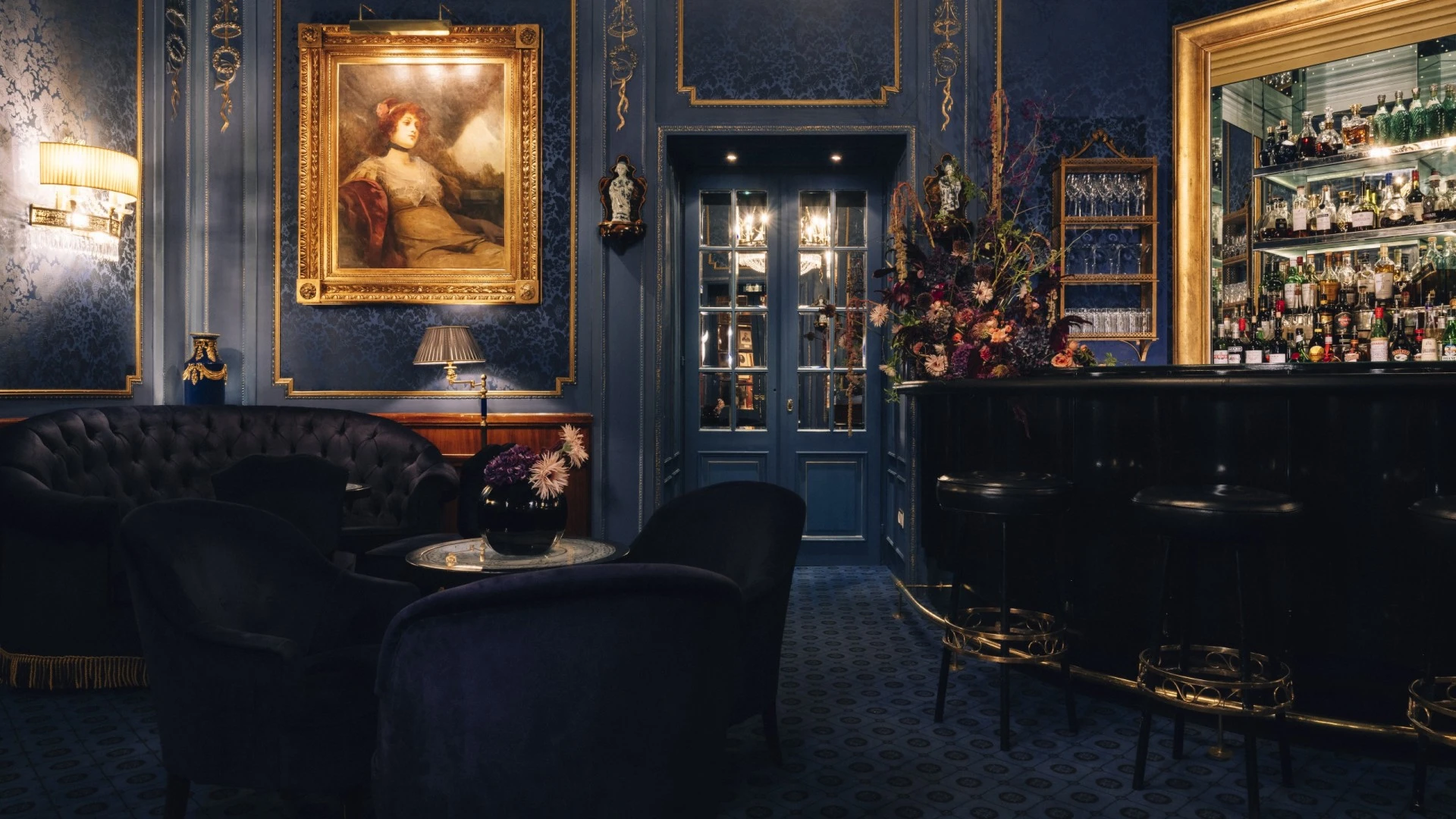 This image offers a full view of the Blaue Bar’s sumptuous interior, with deep blue damask walls, plush velvet seating, a golden-framed mirrored bar, dramatic floral arrangements, and a sparkling chandelier creating an atmosphere of timeless elegance and intimacy.