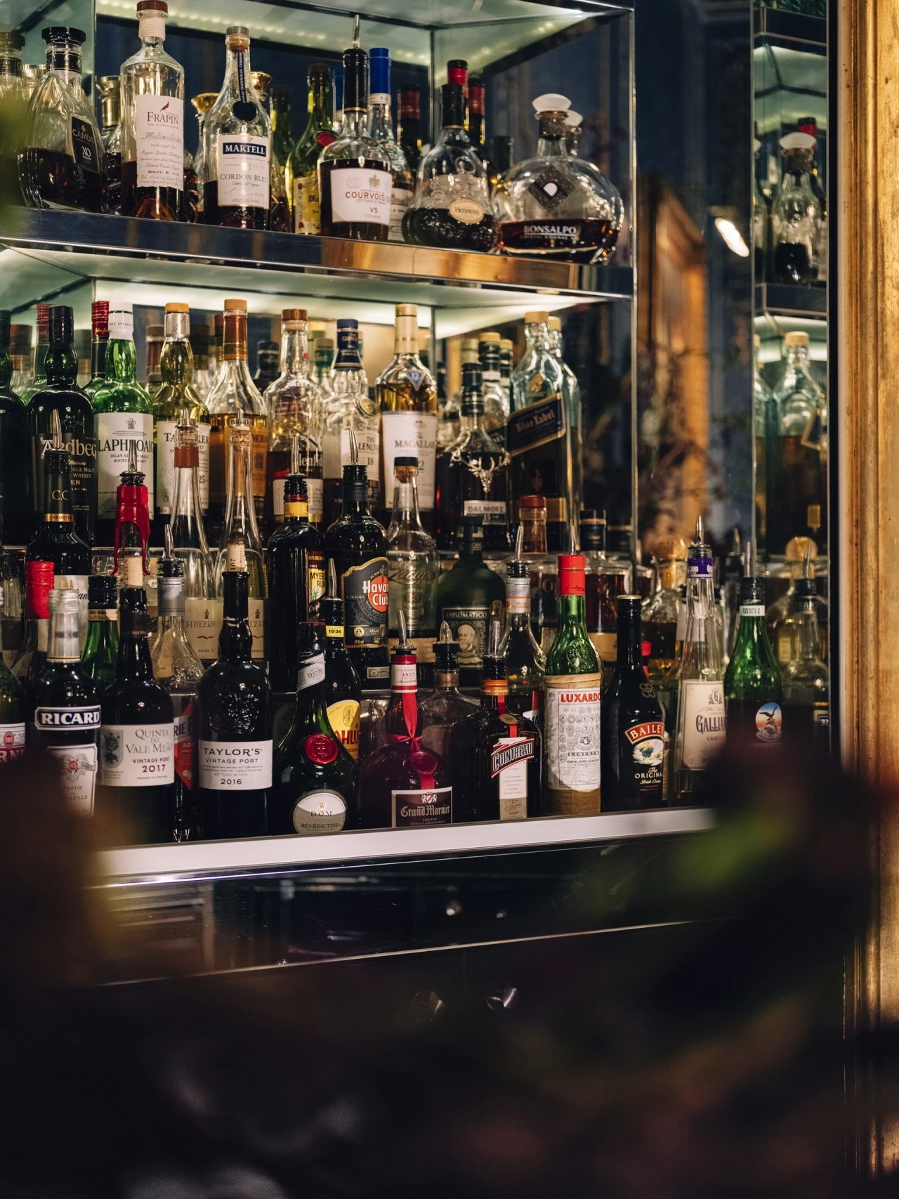 This image shows the lavishly stocked mirrored bar at the Blaue Bar, featuring an exquisite array of premium spirits and liqueurs displayed in a golden-framed cabinet that reflects the bar’s opulent and refined character.