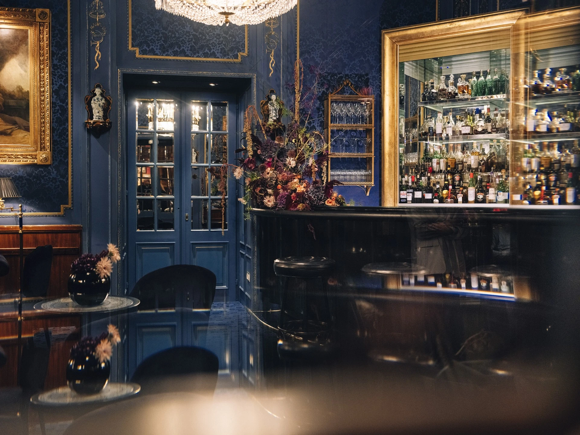 This image captures the refined ambiance of the Blaue Bar, with its signature blue damask wallpaper, ornate gold accents, mirrored bar display, and dramatic floral arrangement creating a sophisticated and intimate setting for cocktails.